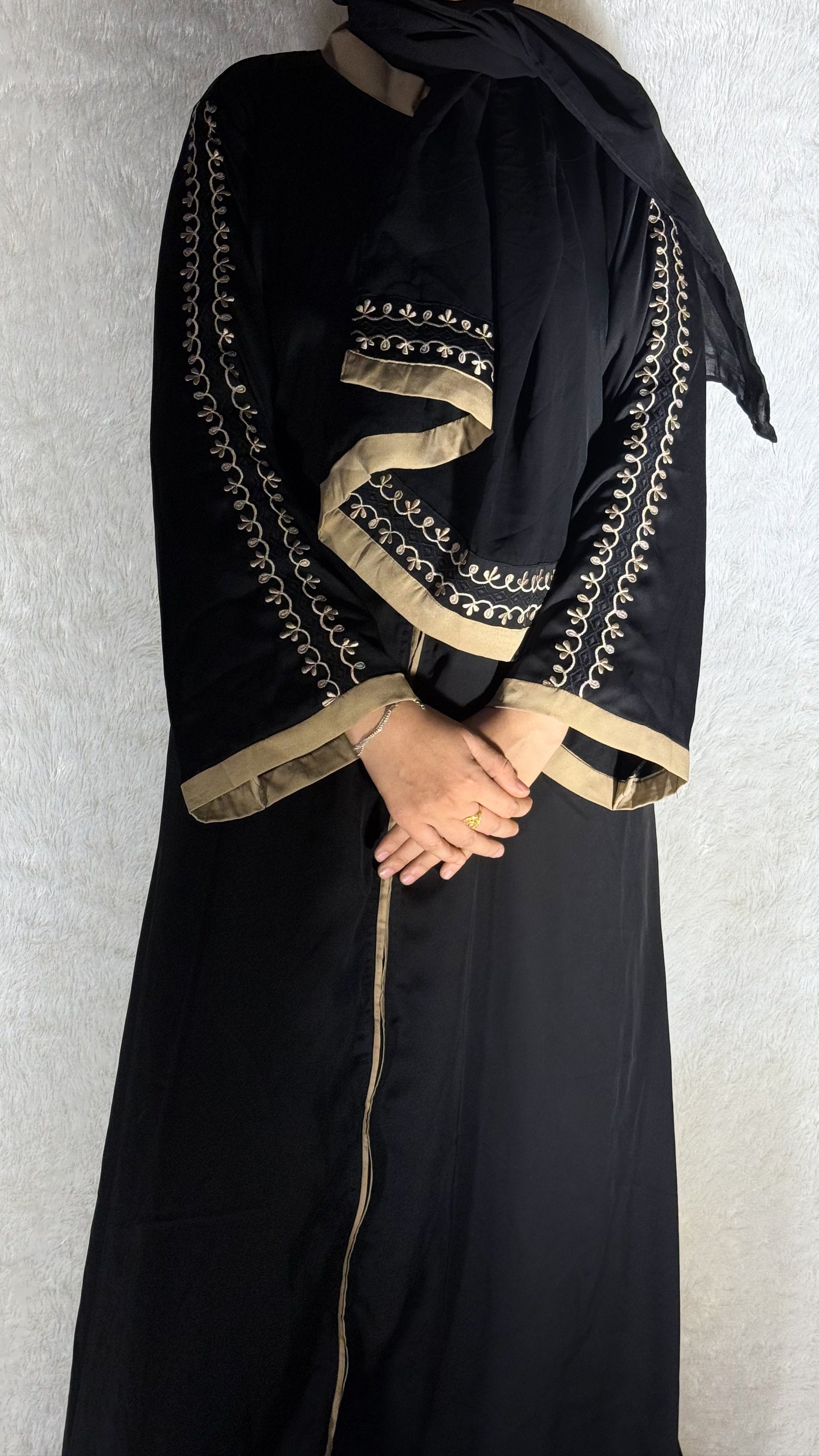 Person wearing a black abaya with gold embroidery on a plain background